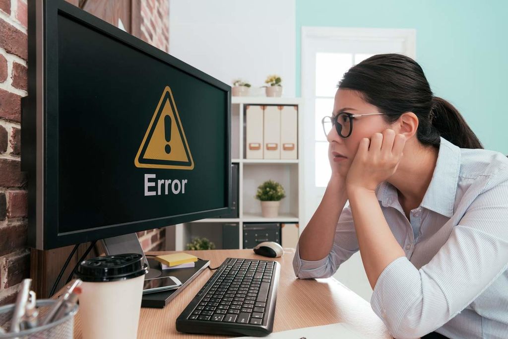 Profissional mulher coloca a mão sobre o rosto em sinal de desaprovação ao ver no monitor do computador um símbolo de alerta de downtime