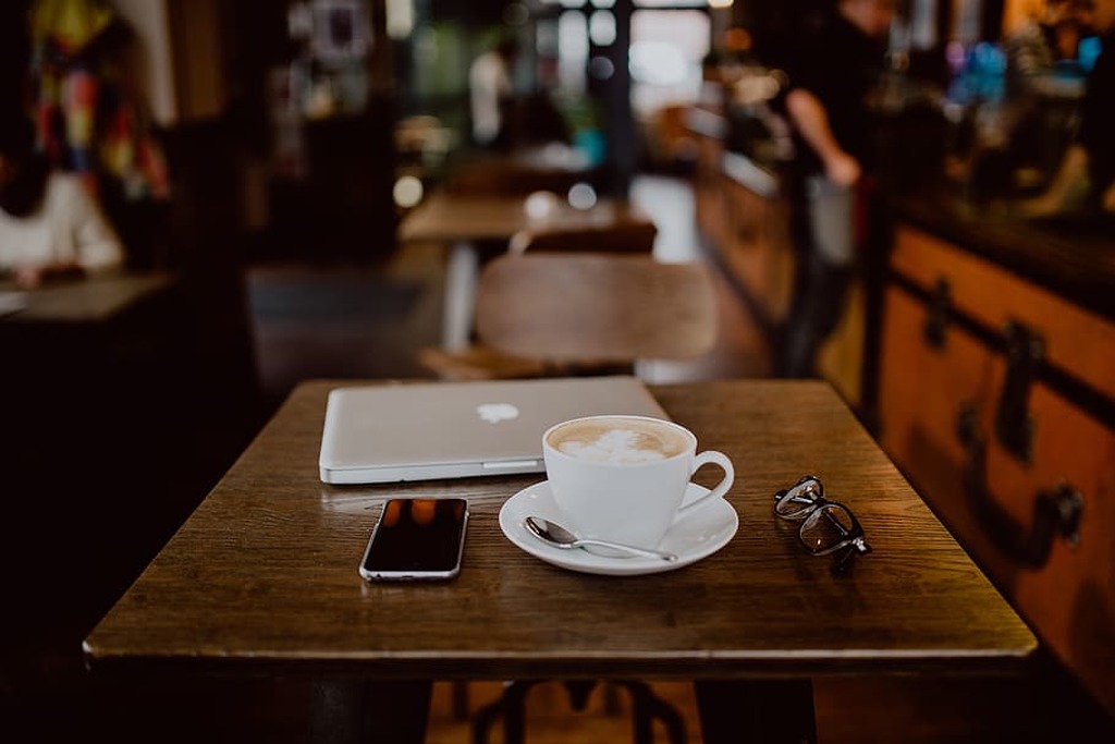 Computador sobre mesa de restaurante. Ao lado, sobre a mesa, um celular e uma xícara de café ilustrando como funciona  a rotina de quem trabalha officeless
